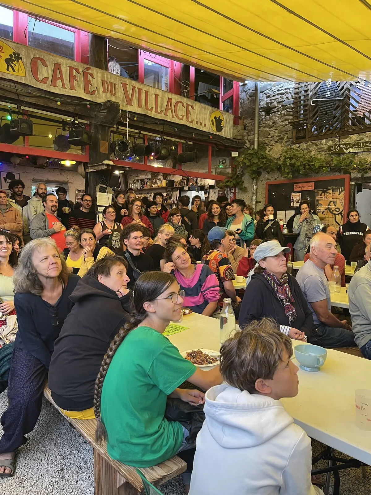La salle du Café pleine, sous l’enseigne Café du Village